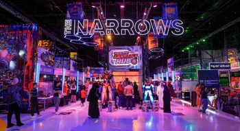 مشهد داخلي لقسم "ذا ناروز" في كوميك كون الشرق الأوسط (MEFCC)، يظهر مجسم روبوت "ميكا" عملاق باللونين البرتقالي والأزرق تحت لوحة نيون مضيئة تحمل عبارة "THE NARROWS"، مع زوار يرتدون ملابس تقليدية وأخرى كاجوال وسط إضاءة نيون درامية.