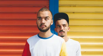 Bigflo et Oli devant un mur coloré, regards croisés et expression sérieuse.