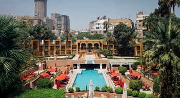 مسبح خارجي كبير بمظلات حمراء وأشجار النخيل داخل منتجع فندقي أخضر، مع إطلالة على مباني المدينة المرتفعة في الخلفية.