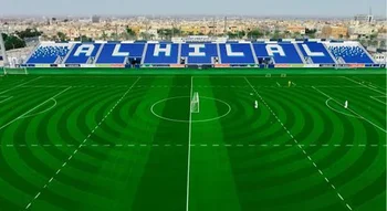 لقطة نهارية لملعب نادي الهلال لكرة القدم، تظهر أرضية عشبية خضراء بتنسيق دائري ومدرجات زرقاء مكتوب عليها اسم النادي باللغة الإنجليزية "AL HILAL" باللون الأبيض، مع ظهور مباني المدينة في الخلفية