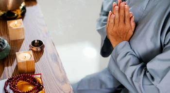لقطة مقربة لشخص يرتدي ثوباً رمادياً يضم يديه معاً في وضعية الصلاة والتأمل أمام طاولة خشبية عليها شموع مضيئة، بخور، ومسبحة حمراء