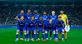 لاعبو فريق الهلال لكرة القدم يرتدون الزي الأزرق الرسمي وحارس المرمى بالزي الأصفر، يقفون في تشكيلة جماعية للالتقاط صورة تذكارية على أرض الملعب قبل بداية المباراة، وتظهر خلفهم مدرجات مليئة بالجماهير تحت الأضواء الكاشفة.