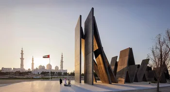 نصب واحة الكرامة التذكاري في أبوظبي المكون من ألواح معدنية ضخمة متكئة على بعضها، مع ظهور جامع الشيخ زايد الكبير وعلم الإمارات في الخلفية وقت الغروب.