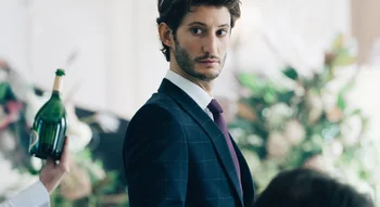 Pierre Niney en costume-cravate regardant par-dessus son épaule avec intensité.