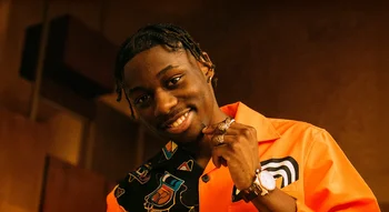 Portrait souriant de Tiakola en chemise orange décorée, posant avec des bagues.