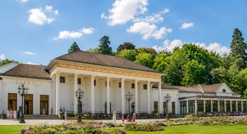 Sommeransicht der Spielbank Baden-Baden im Kurhaus mit neoklassizistischer Fassade, Säulen, blühenden Beeten und Spaziergängern.