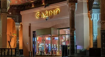 مدخل فخم لكازينو مصمم على الطراز المعماري المغربي التقليدي المميز، بأعمدة مزخرفة وأسقف فنية. تظهر في الواجهة لافتة مضيئة باسم "CASINO" فوق مدخل زجاجي دوار. يمكن رؤية آلات القمار المضاءة داخل المبنى كهرومغناطيسياً. يوجد رجلان يقفان بالقرب من المدخل، والإضا
