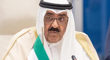 صورة لمسؤول خليجي رفيع المستوى يرتدي الزي التقليدي (الغترة البيضاء، العقال، والبشت) ونظارات طبية، يجلس في مؤتمر أو اجتماع رسمي مع وجود ميكروفون وجزء من علم دولة الكويت أمامه.