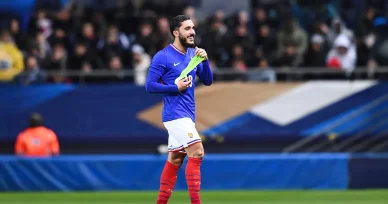 Rayan Cherki marche sur le terrain avec le maillot bleu français. Rayan Cherki sourit en tenant un brassard avec l’équipe de France.