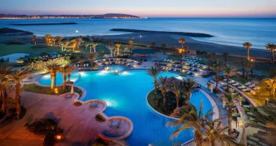 إطلالة بانورامية لمنتجع شاطئي فاخر يتميز بحمامات سباحة مضيئة وأشجار النخيل عند الغسق.