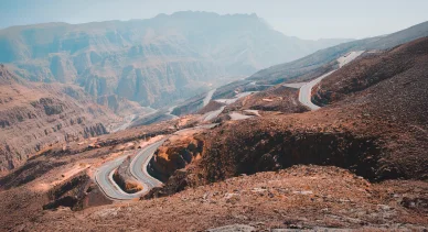طريق أسفلتي متعرج يمر عبر جبال صخرية وعرة في مشهد طبيعي صحراوي.