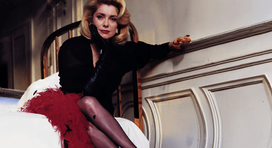 Catherine Deneuve assise avec élégance dans une chambre, portant une robe noire et une jupe rouge à plumes.