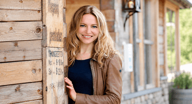 Diana Staehly lächelt in einer braunen Lederjacke vor einer Holzhütte mit Öffnungszeiten-Schild