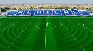 لقطة نهارية لملعب نادي الهلال لكرة القدم، تظهر أرضية عشبية خضراء بتنسيق دائري ومدرجات زرقاء مكتوب عليها اسم النادي باللغة الإنجليزية "AL HILAL" باللون الأبيض، مع ظهور مباني المدينة في الخلفية