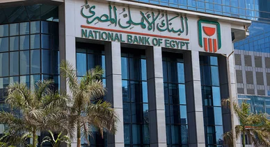الواجهة الزجاجية الحديثة لمبنى البنك الأهلي المصري مع علامة البنك والاسم باللغتين العربية والإنجليزية، وتظهر أشجار النخيل في المقدمة.
