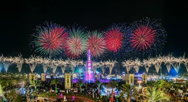 حشد كبير من الزوار يشاهدون عرضاً مبهراً للألعاب النارية الملونة تتلألأ في سماء الليل فوق هيكل مضاء في ساحة المهرجان.