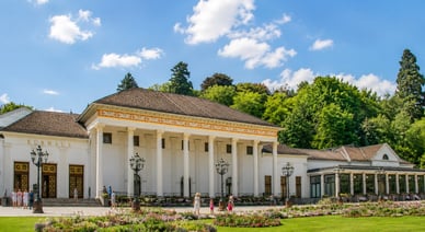 Sommeransicht der Spielbank Baden-Baden im Kurhaus mit neoklassizistischer Fassade, Säulen, blühenden Beeten und Spaziergängern.