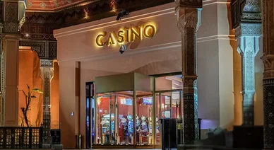 مدخل فخم لكازينو مصمم على الطراز المعماري المغربي التقليدي المميز، بأعمدة مزخرفة وأسقف فنية. تظهر في الواجهة لافتة مضيئة باسم "CASINO" فوق مدخل زجاجي دوار. يمكن رؤية آلات القمار المضاءة داخل المبنى كهرومغناطيسياً. يوجد رجلان يقفان بالقرب من المدخل، والإضا