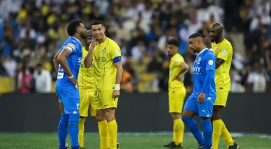 كريستيانو رونالدو بقميص نادي النصر الأصفر يبتسم ويتحدث مع أحد لاعبي نادي الهلال بالقميص الأزرق أثناء توقف اللعب في مباراة كرة قدم، مع ظهور لاعبين آخرين من كلا الفريقين ومدرجات ممتلئة بالمشجعين في الخلفية ضبابية.
