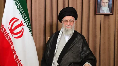 المرشد الأعلى للجمهورية الإسلامية الإيرانية، آية الله علي خامنئي، يجلس بالزي الديني الأسود بجوار علم إيران، وتظهر في الخلفية صورة للإمام الخميني