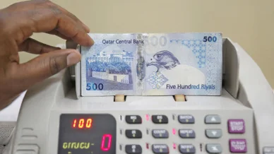 آلة إلكترونية لعد الأوراق النقدية القطرية يد تضع ورقة نقدية من فئة 500 ريال قطري، يظهر عليها رسم لصقر ومبنى مصرف قطر المركزي، داخل آلة إلكترونية لعد النقود.