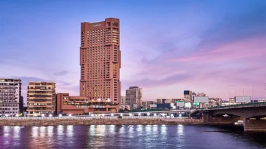 فندق رمسيس هيلتون الشاهق يطل على نهر النيل في القاهرة وقت الغسق، مع سماء ملونة بتدرجات الأرجواني والوردي وجسر يمر فوق النهر