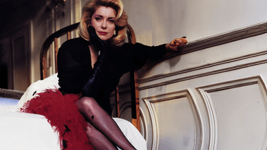 Catherine Deneuve assise avec élégance dans une chambre, portant une robe noire et une jupe rouge à plumes.