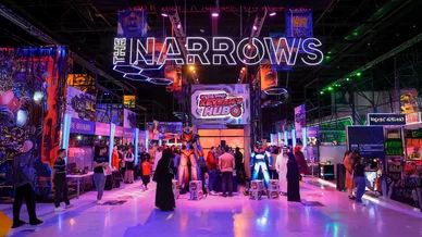 مشهد داخلي لقسم "ذا ناروز" في كوميك كون الشرق الأوسط (MEFCC)، يظهر مجسم روبوت "ميكا" عملاق باللونين البرتقالي والأزرق تحت لوحة نيون مضيئة تحمل عبارة "THE NARROWS"، مع زوار يرتدون ملابس تقليدية وأخرى كاجوال وسط إضاءة نيون درامية.