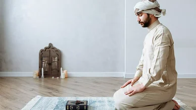 رجل عربي بملابس تقليدية يجلس بخشوع على سجادة صلاة، وأمامه نسخة من القرآن الكريم ومسبحة.