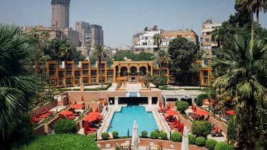 مسبح خارجي كبير بمظلات حمراء وأشجار النخيل داخل منتجع فندقي أخضر، مع إطلالة على مباني المدينة المرتفعة في الخلفية.