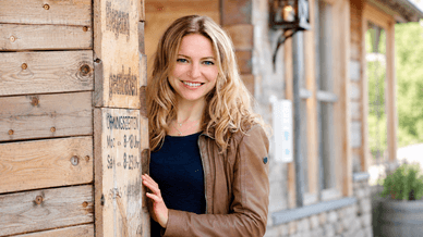 Diana Staehly lächelt in einer braunen Lederjacke vor einer Holzhütte mit Öffnungszeiten-Schild