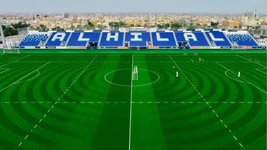 ملعب نادي الهلال: جاهزية وأناقة في معقل الزعيم لقطة نهارية لملعب نادي الهلال لكرة القدم، تظهر أرضية عشبية خضراء بتنسيق دائري ومدرجات زرقاء مكتوب عليها اسم النادي باللغة الإنجليزية "AL HILAL" باللون الأبيض، مع ظهور مباني المدينة في الخلفية