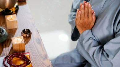 التأمل والسكينة: طقوس الصلاة والتركيز الذهني لقطة مقربة لشخص يرتدي ثوباً رمادياً يضم يديه معاً في وضعية الصلاة والتأمل أمام طاولة خشبية عليها شموع مضيئة، بخور، ومسبحة حمراء