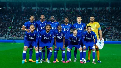 لاعبو فريق الهلال لكرة القدم يرتدون الزي الأزرق الرسمي وحارس المرمى بالزي الأصفر، يقفون في تشكيلة جماعية للالتقاط صورة تذكارية على أرض الملعب قبل بداية المباراة، وتظهر خلفهم مدرجات مليئة بالجماهير تحت الأضواء الكاشفة.