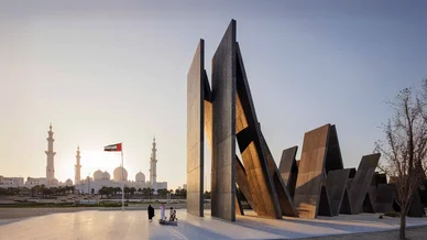 نصب واحة الكرامة التذكاري في أبوظبي المكون من ألواح معدنية ضخمة متكئة على بعضها، مع ظهور جامع الشيخ زايد الكبير وعلم الإمارات في الخلفية وقت الغروب.