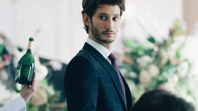 Pierre Niney en costume-cravate regardant par-dessus son épaule avec intensité.