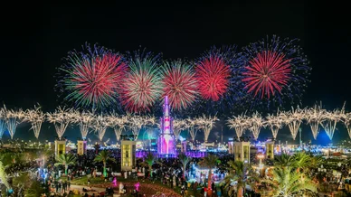 حشد كبير من الزوار يشاهدون عرضاً مبهراً للألعاب النارية الملونة تتلألأ في سماء الليل فوق هيكل مضاء في ساحة المهرجان.