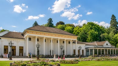 Sommeransicht der Spielbank Baden-Baden im Kurhaus mit neoklassizistischer Fassade, Säulen, blühenden Beeten und Spaziergängern.