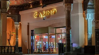 مدخل فخم لكازينو مصمم على الطراز المعماري المغربي التقليدي المميز، بأعمدة مزخرفة وأسقف فنية. تظهر في الواجهة لافتة مضيئة باسم "CASINO" فوق مدخل زجاجي دوار. يمكن رؤية آلات القمار المضاءة داخل المبنى كهرومغناطيسياً. يوجد رجلان يقفان بالقرب من المدخل، والإضا