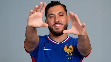Rayan Cherki pose en portant le maillot bleu de la France. Rayan Cherki souriant avec le maillot de l’équipe de France.