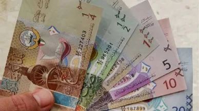 يد تمسك بمروحة من الأوراق النقدية للدينار الكويتي بمختلف فئاتها، بدءاً من ربع ونصف دينار، وصولاً إلى 1 و 5 و 10 و 20 ديناراً، مما يبرز الرمز الوطني وتنوع الألوان والتصاميم