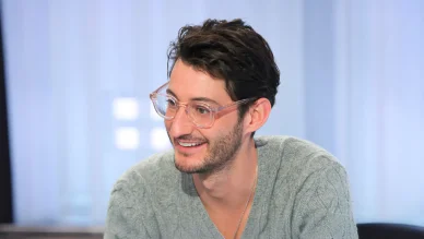 Portrait de Pierre Niney portant des lunettes à monture transparente et souriant durant une interview.