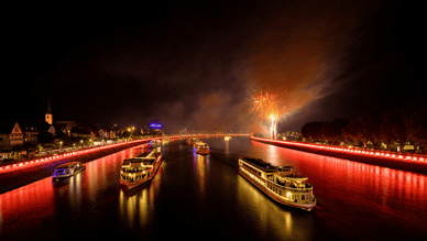 „Rhein in Flammen“ bei Nacht – mehrere Ausflugsschiffe fahren auf dem Rhein zwischen den beleuchteten Ufern hindurch, während am Horizont ein Feuerwerk zu sehen ist