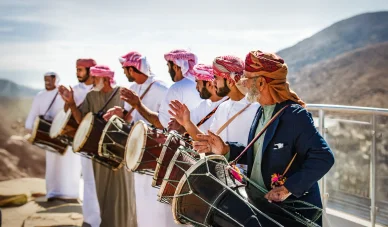 فرقة عمانية تؤدي رقصة شعبية مع القرع على طبول كبيرة في مشهد جبلي مفتوح.