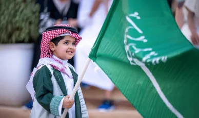 فرحة طفل سعودي باليوم الوطني مع علم المملكة طفل سعودي بملابس تقليدية يحمل علم السعودية مبتسماً في احتفال.