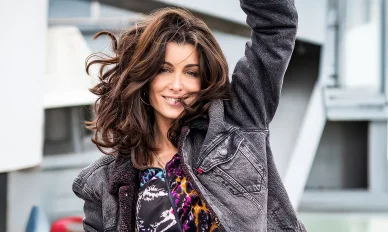 Une photo joyeuse de la chanteuse Jenifer prise en extérieur. Jenifer souriante et pleine de vie avec une veste en jean et les cheveux au vent.