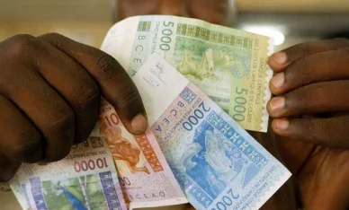 التعاملات النقدية وقيمة العملة في دول البنك المركزي لدول غرب أفريقيا فرنك غرب أفريقيا (CFA Franc): يدان تمسكان بأوراق نقدية من فئات مختلفة مثل 5000 و 2000 و 10000.