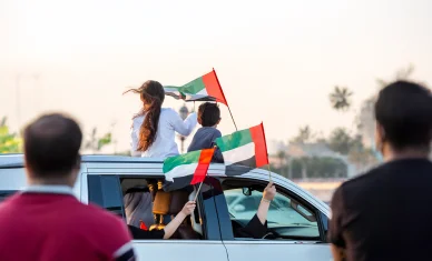 لقطة خارجية من الخلف لأطفال يحتفلان على سطح سيارة مكشوفة، يلوحان بعلم الإمارات، وسط حشد خارجي مشوش وأجواء احتفالية.