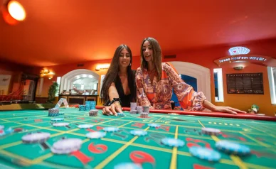 امرأتان تضعان الرقائق على طاولة روليت خضراء كلاسيكية أثناء اللعب داخل صالة كازينو ذات جدران حمراء.