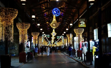 ممر داخل سوق المباركية التقليدي في الكويت ليلاً، مزين بثريات متلألئة وأضواء زينة مع أشخاص يتجولون بين المتاجر.